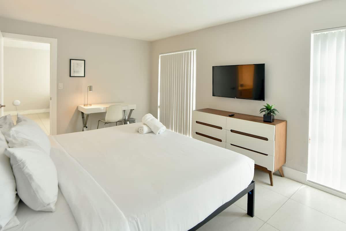 A modern hotel room with a large bed, a desk, a chair, a TV, and a window with blinds.