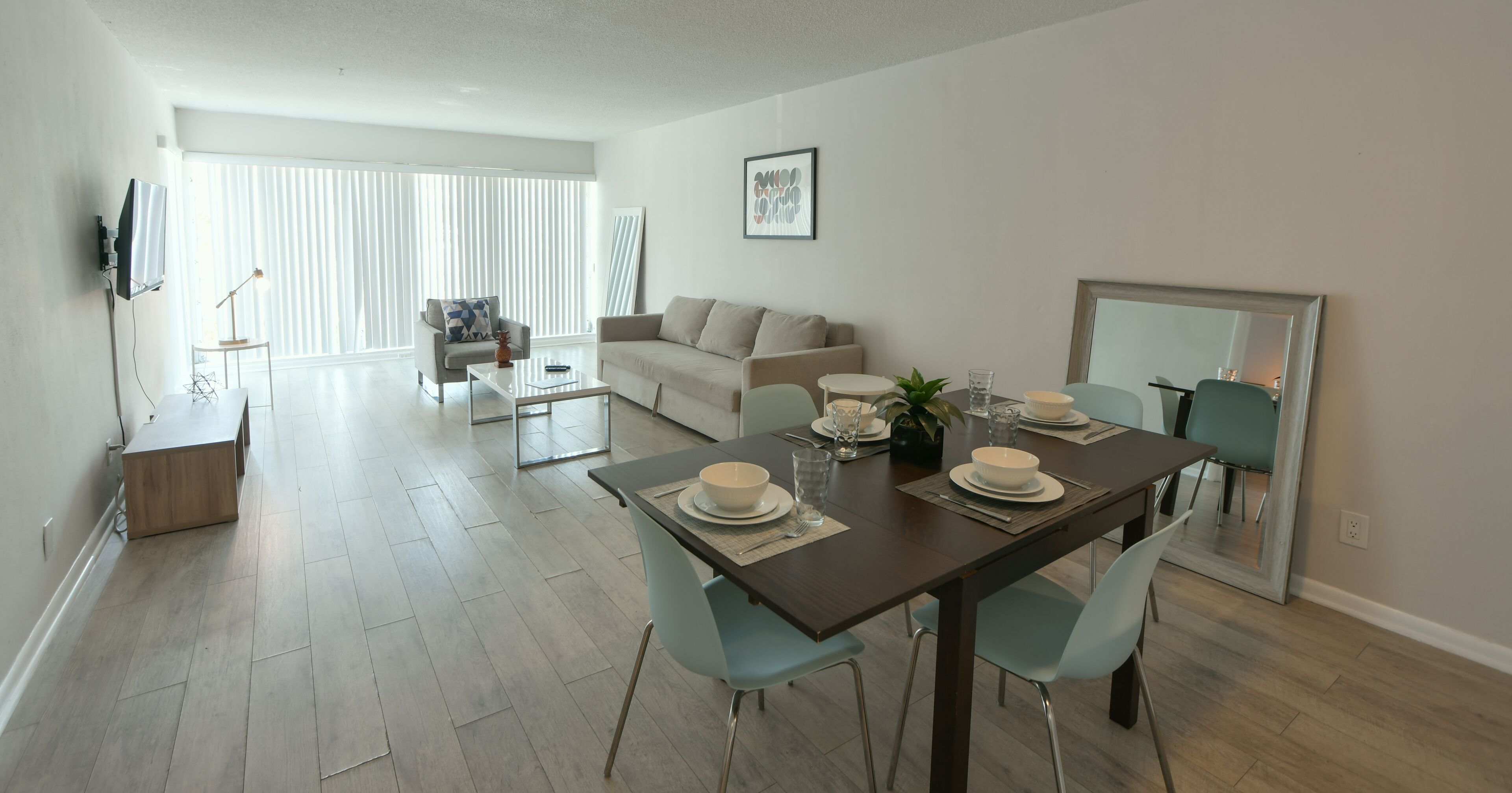 A modern dining area with a wooden table set for four, light blue chairs, and a large mirror on the wall.