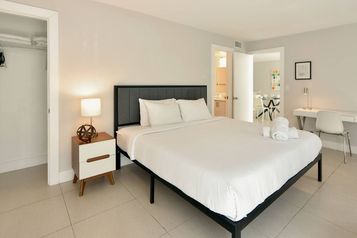 A modern hotel room with a large bed, bedside tables, a desk, and a bathroom visible through an open door.