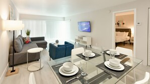 Un salon moderne avec une table à manger en verre pour quatre personnes, un canapé gris, un fauteuil bleu et un téléviseur fixé au mur.