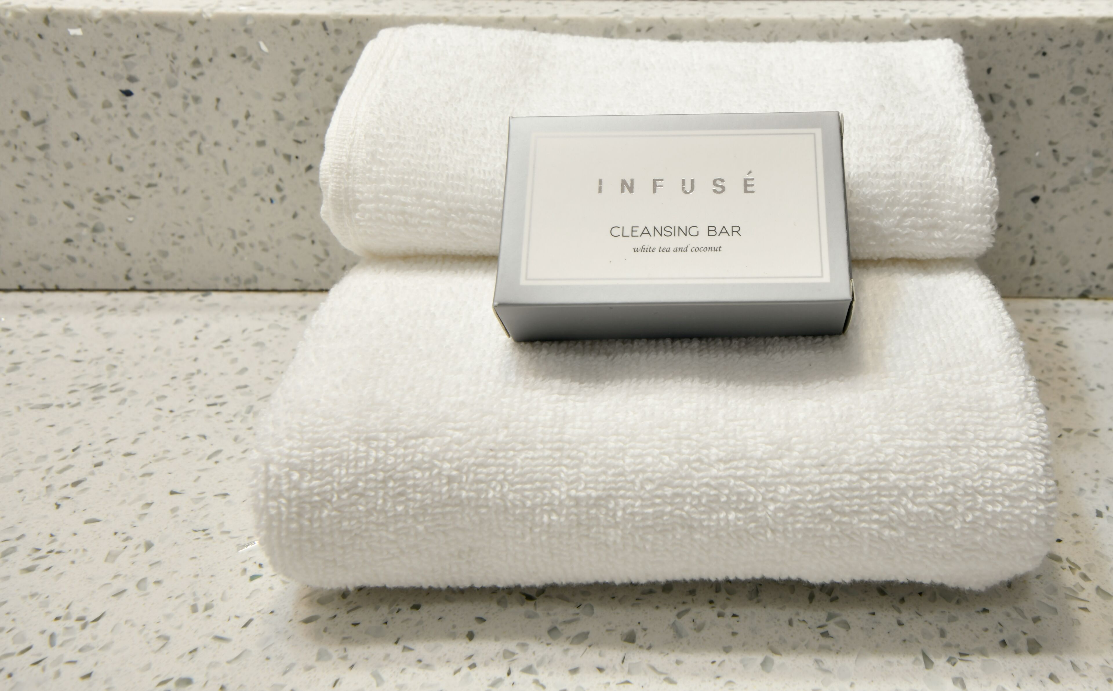 Una toalla blanca con una caja etiquetada como “INFUSE CLEANSING BAR” sobre una encimera con motas.