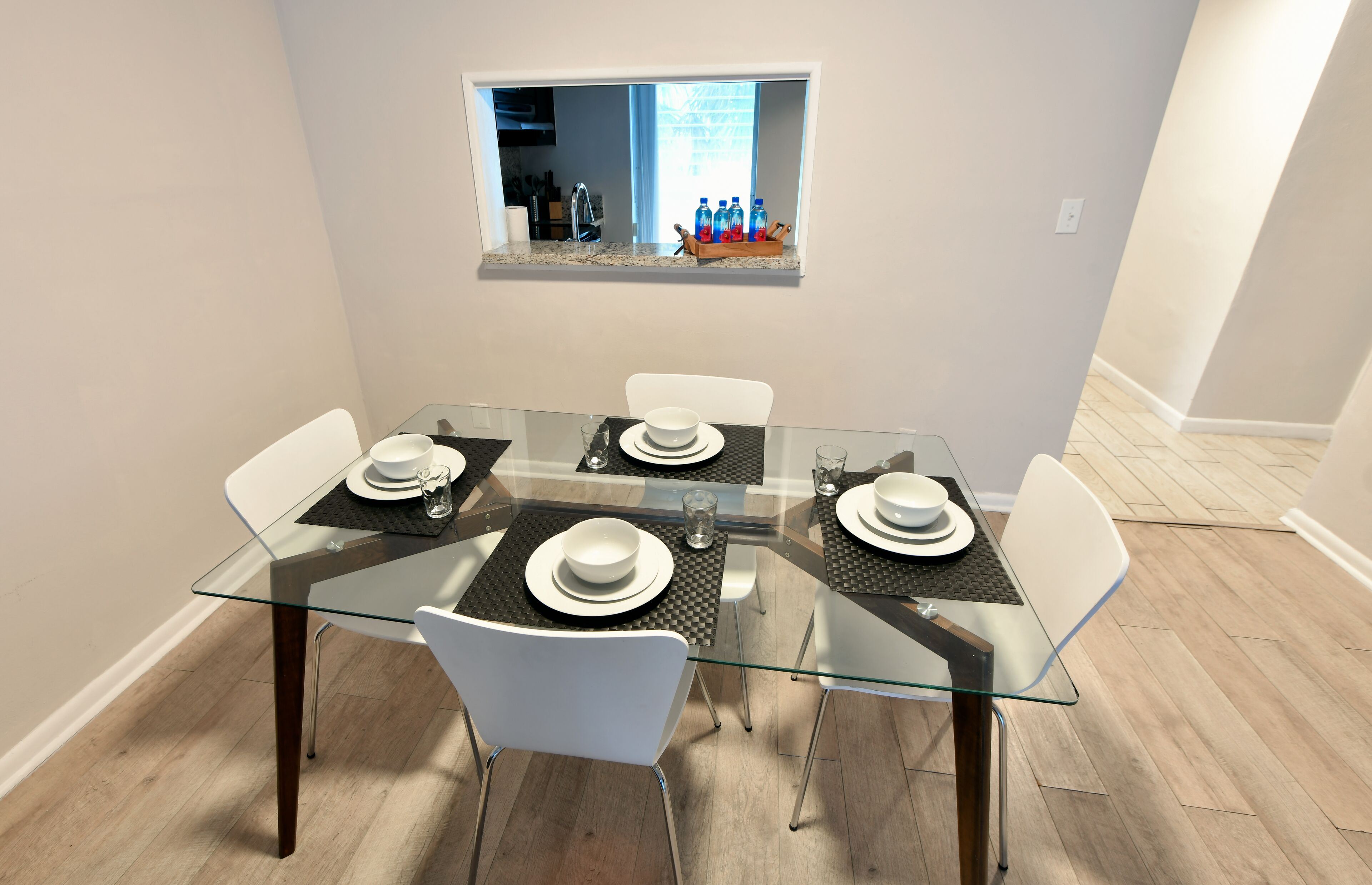 Un coin repas avec une table en verre, des chaises blanches et une fenêtre donnant sur la cuisine.