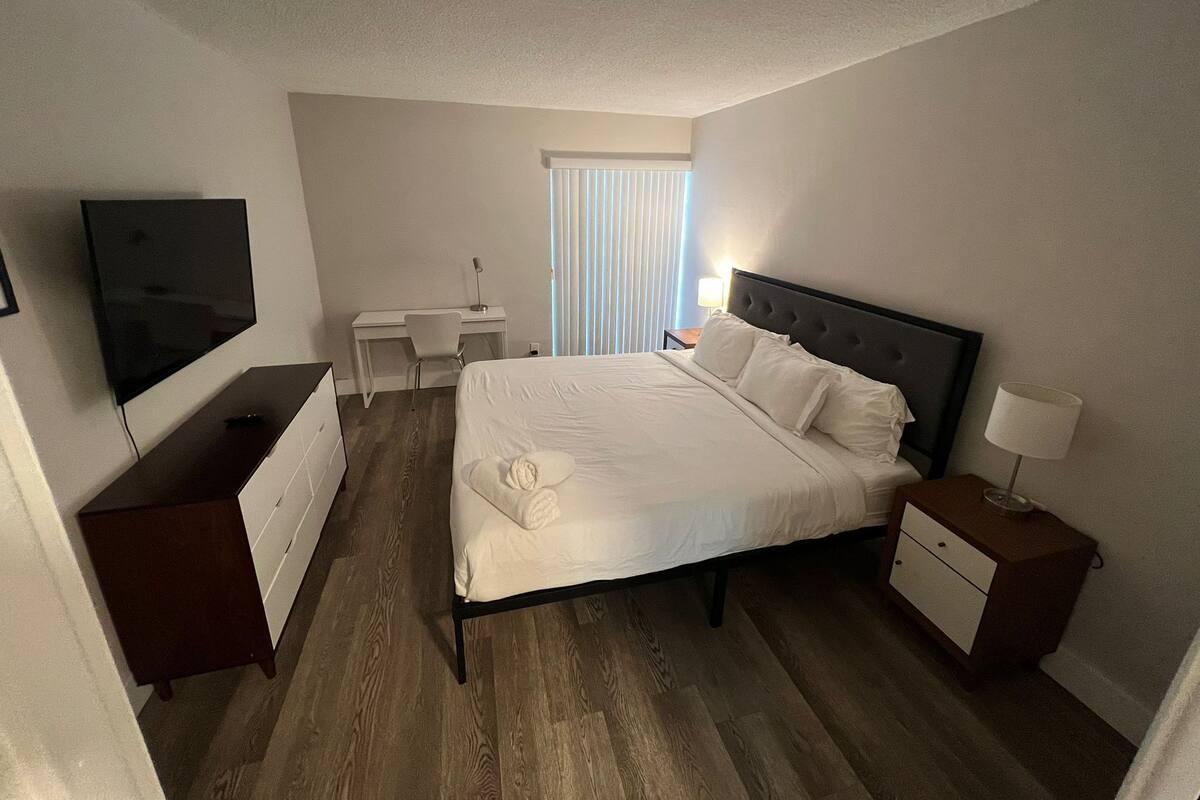 A hotel room with a bed, a desk, a TV, and a window with blinds.