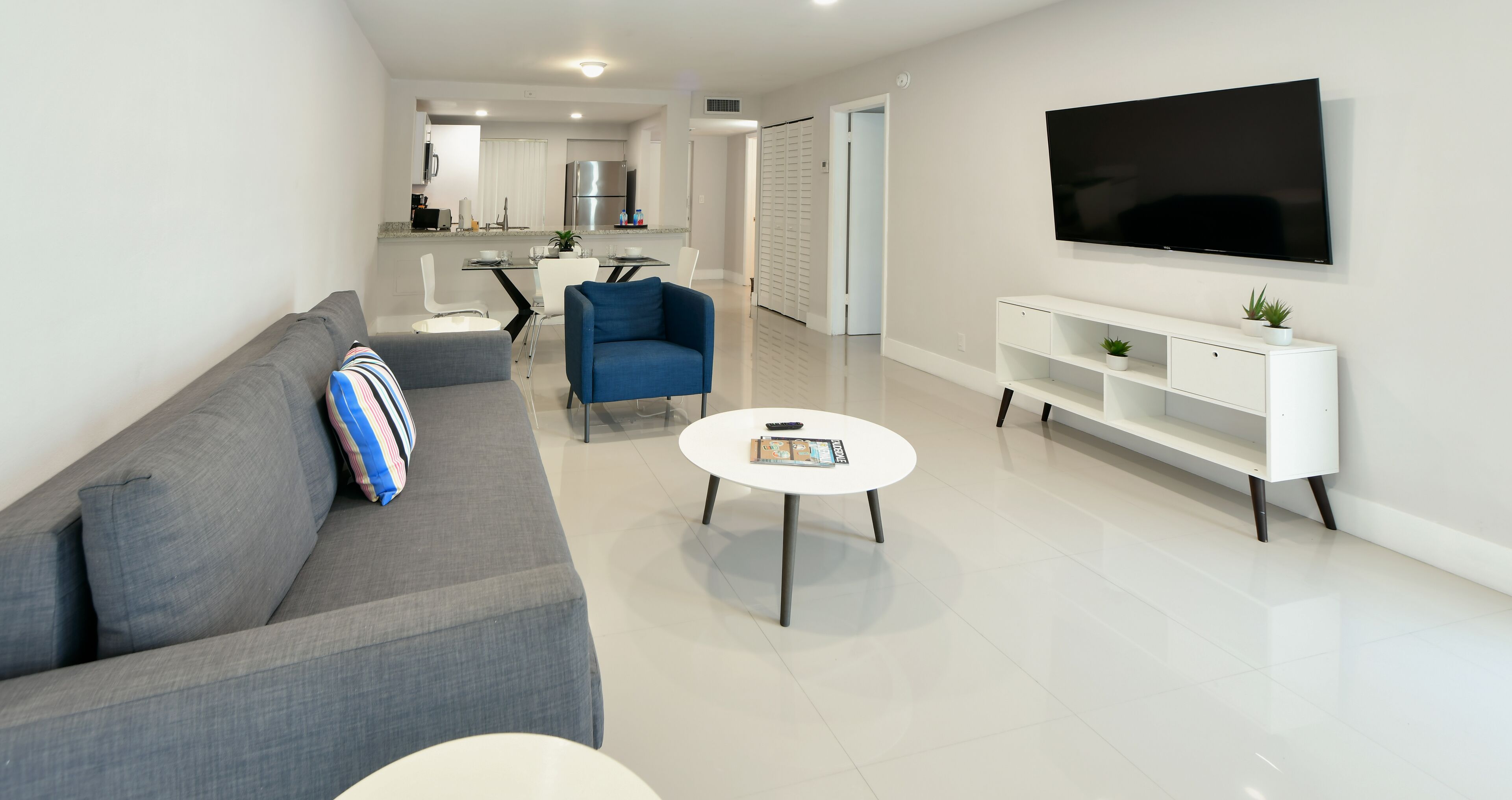 A modern living room with a grey sofa, a blue armchair, a white coffee table, and a flat-screen TV mounted on the wall.