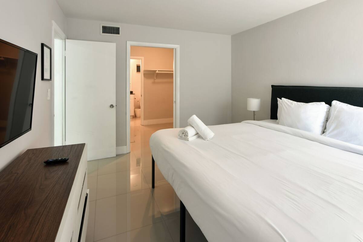 A hotel room with a bed, a TV, a desk, and a bathroom visible through an open door.