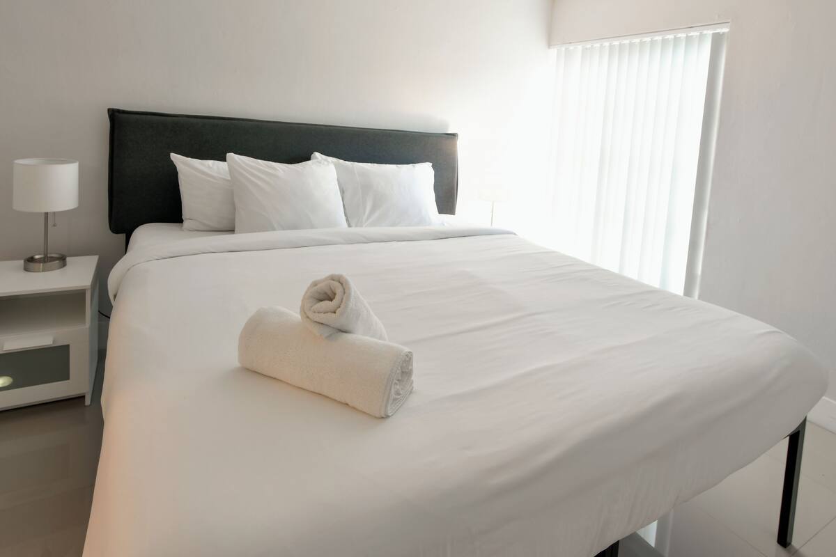 A neatly made bed with white linens and pillows, a rolled towel, a bedside table with a lamp, and a window with blinds.