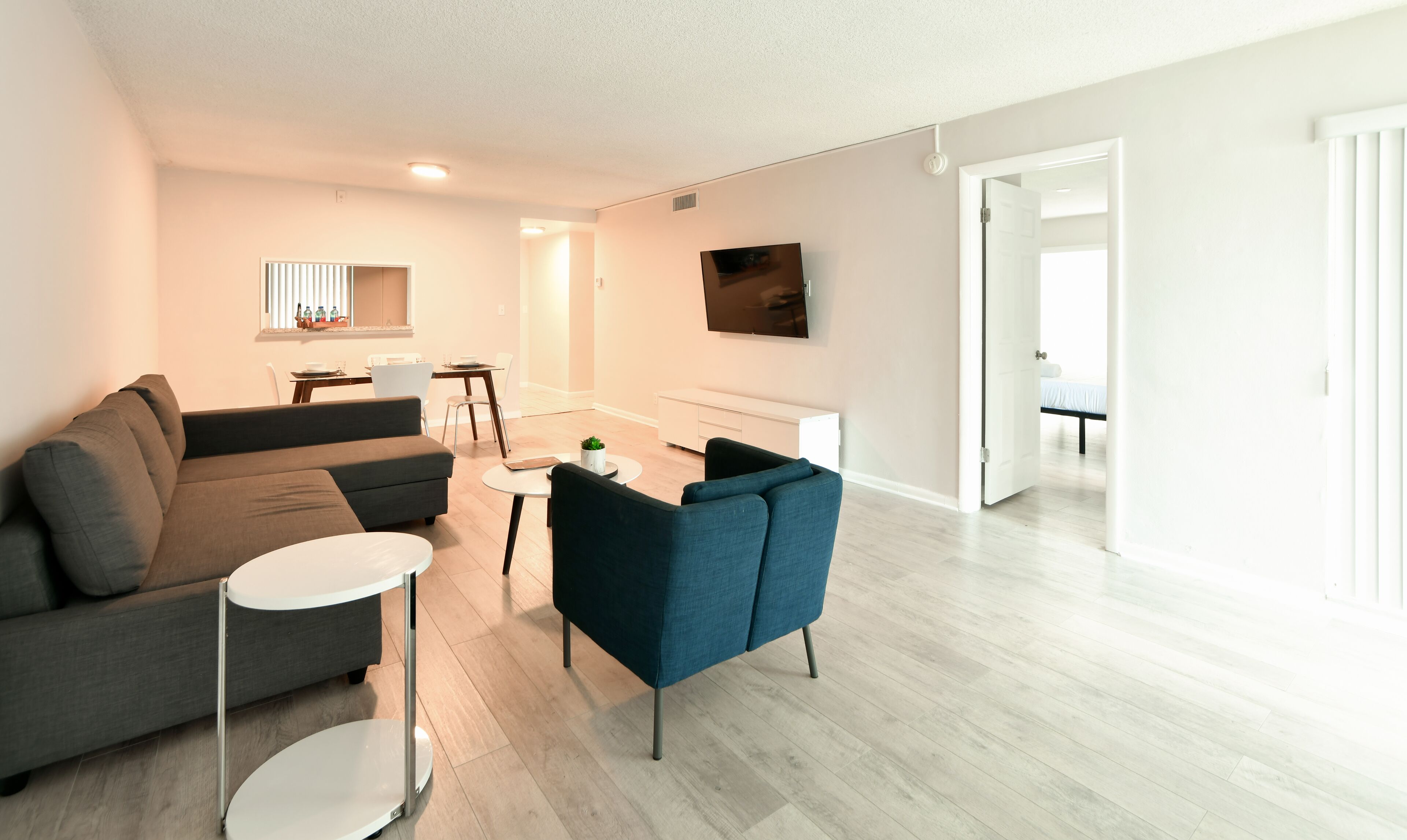Una sala moderna con un sofá gris, un sillón azul y una mesita blanca. Hay una zona de comedor con una mesa y sillas, y un televisor montado en la pared.
