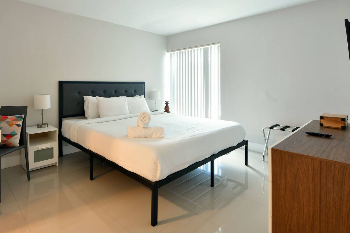 A modern hotel room with a neatly made bed, a small desk, a chair, and a bedside table with a lamp.