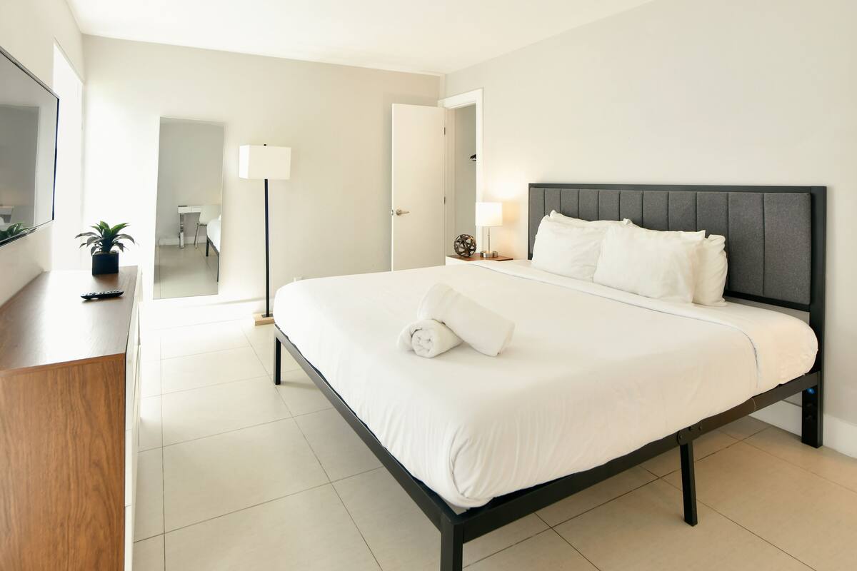 A modern hotel room with a large bed, a flat-screen TV mounted on the wall, a bedside table with a lamp, and a view of the bathroom through an open door.