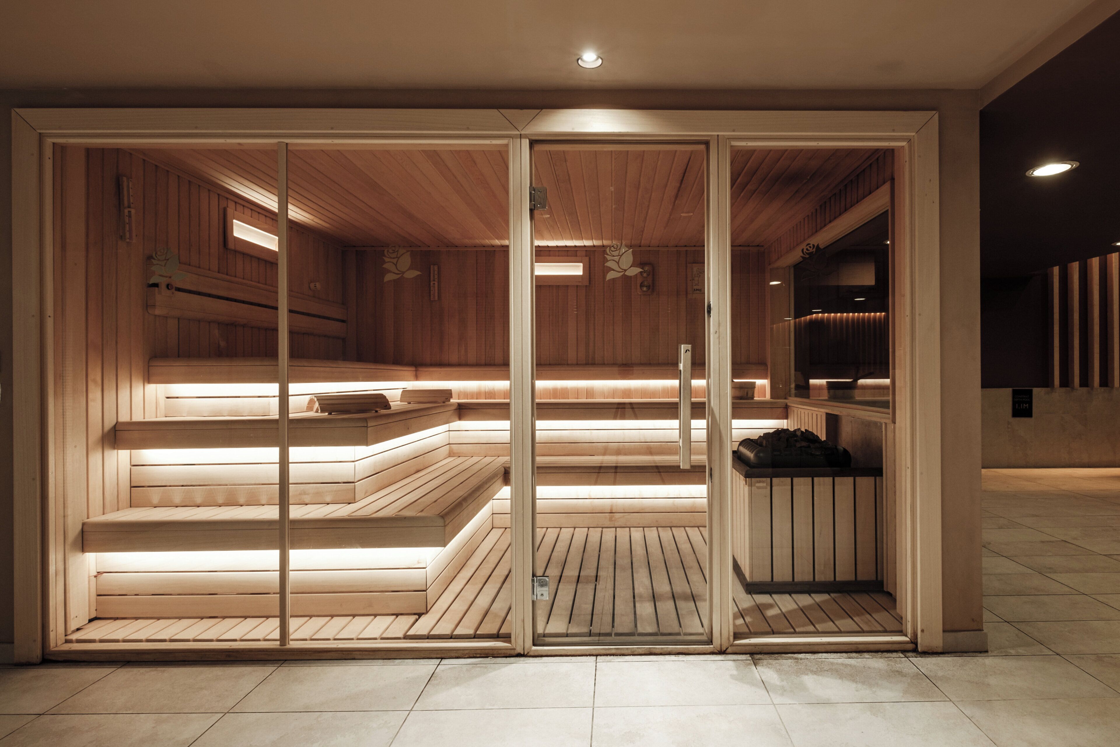 Una sauna con bancos de madera y suelo de baldosas, con una puerta de cristal y una luz en el techo.