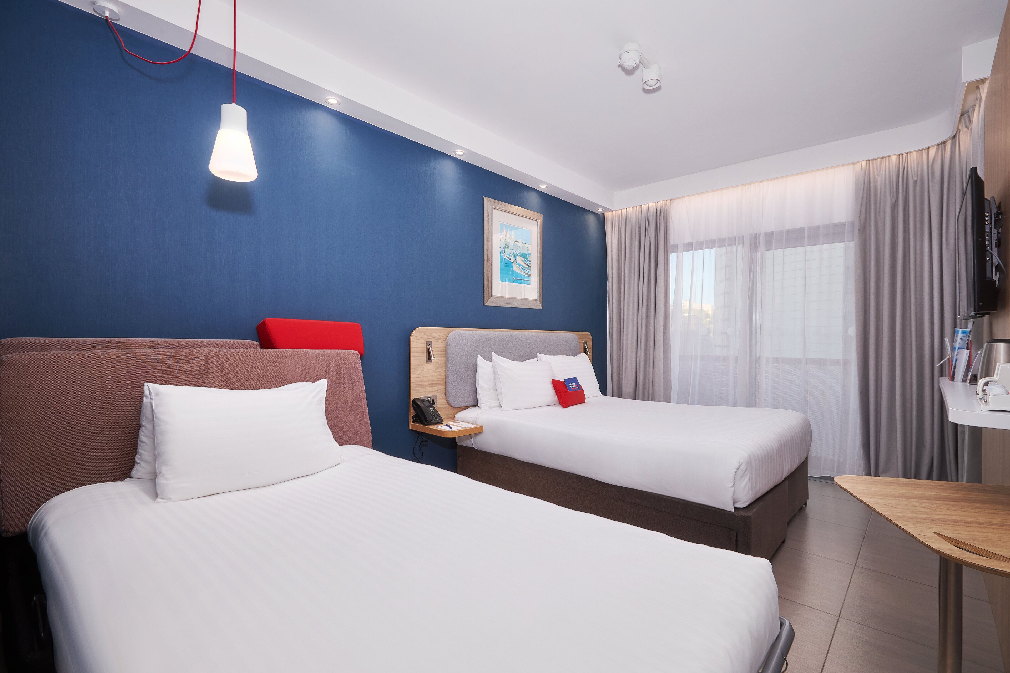 A hotel room with two beds, a desk, a TV, and a window with curtains.