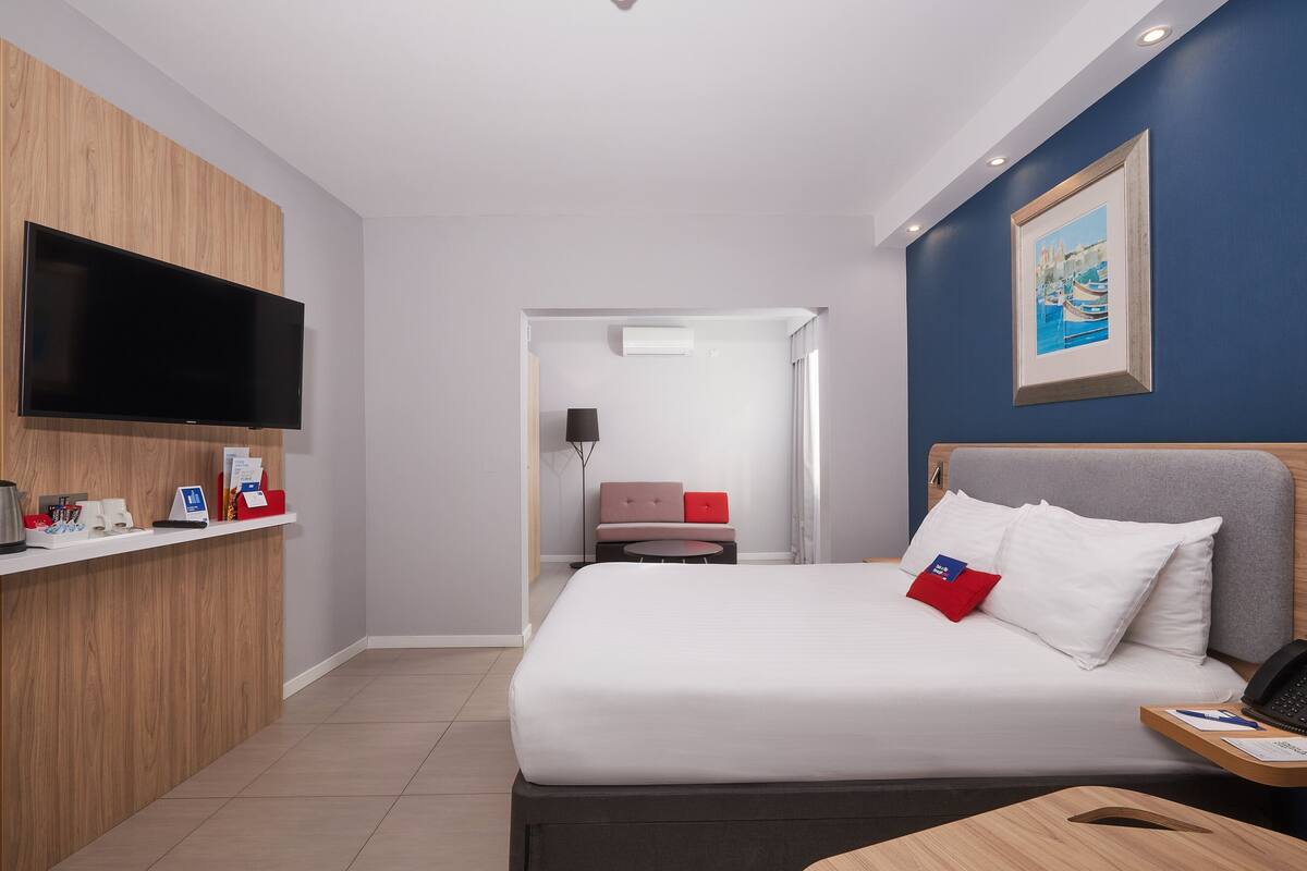 A hotel room with a bed, a desk with a flat-screen TV, a framed picture on the wall, and a door leading to another room.