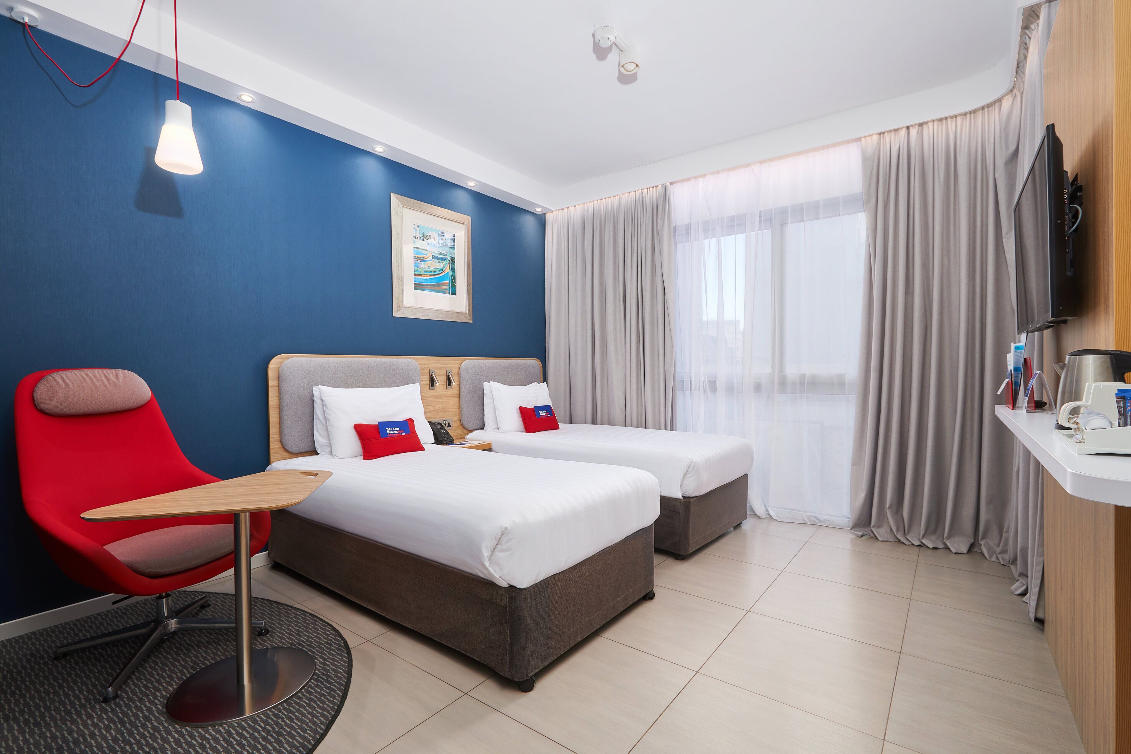 A hotel room with two beds, a red chair, a desk, a painting on the wall, and a TV on the wall.