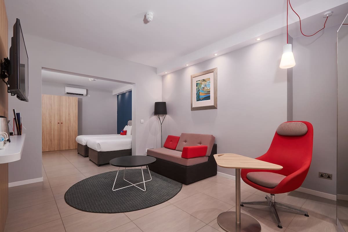 A modern hotel room with a bed, a sofa, a red chair, a small table, and a TV mounted on the wall.