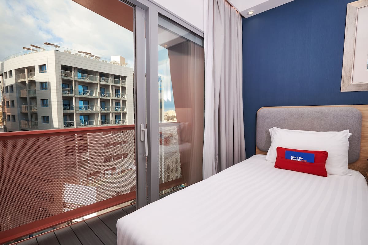 A hotel room with a balcony, a bed with a red pillow, and a view of a building across the street.