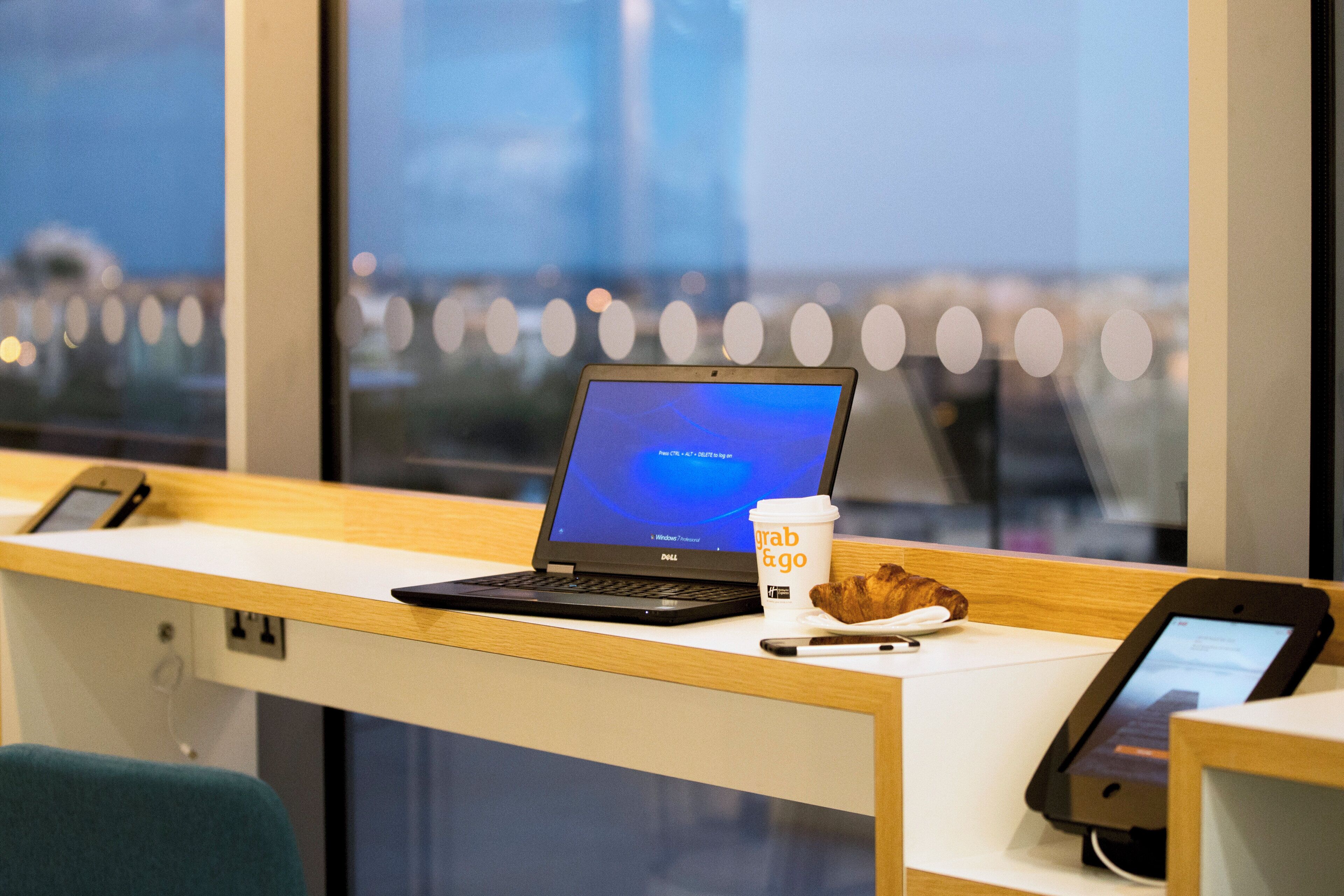 Una laptop sobre un escritorio con una taza de café y un croissant, un teléfono inteligente, y una ventana con vistas a edificios.
