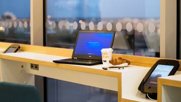 Una laptop sobre un escritorio con una taza de café y un croissant, un teléfono inteligente, y una ventana con vistas a edificios.