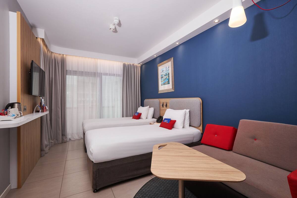 A hotel room with two beds, a sofa, a TV, a desk, and a painting on the wall.