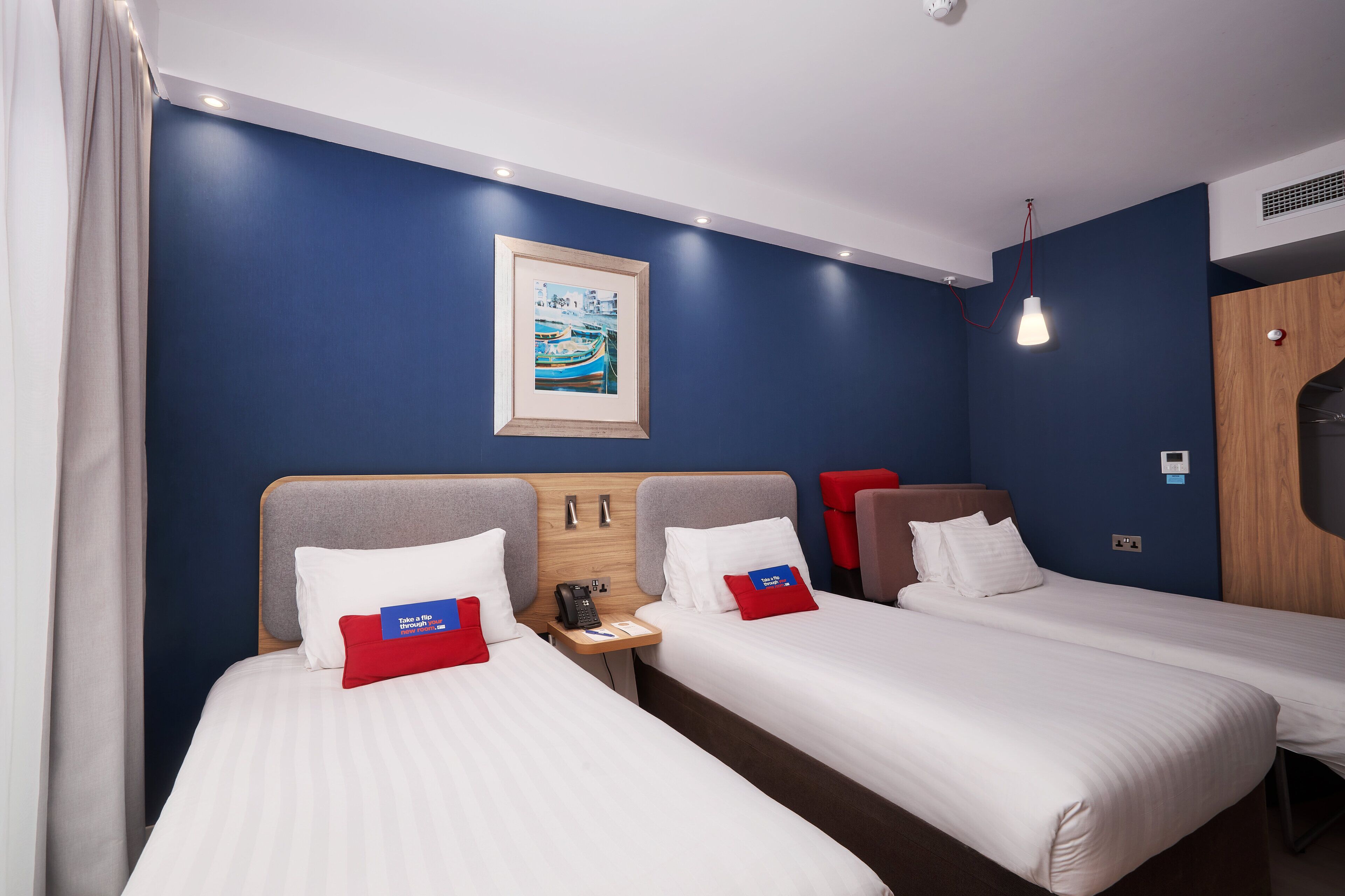 A hotel room with two single beds, a wooden headboard, a nightstand with a phone, and a painting on the wall.