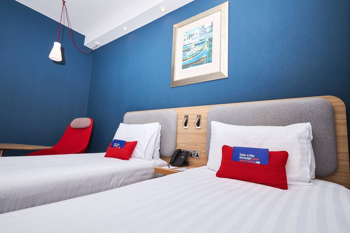 A hotel room with two beds, a red chair, a desk, and a framed picture on the wall.