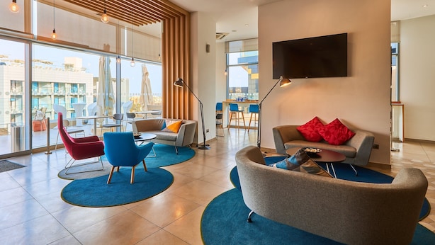 A modern hotel lobby with a large window, a flat-screen TV, and a seating area with a sofa and chairs. - Holiday Inn Express Malta by IHG (St. Julian's)