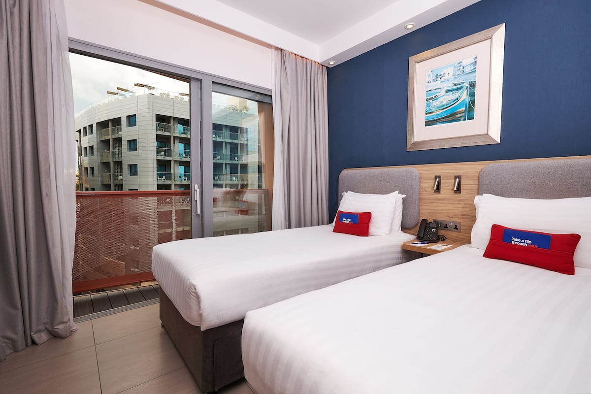 A hotel room with two beds, a large window, a framed picture on the wall, and a balcony with a view of a building.