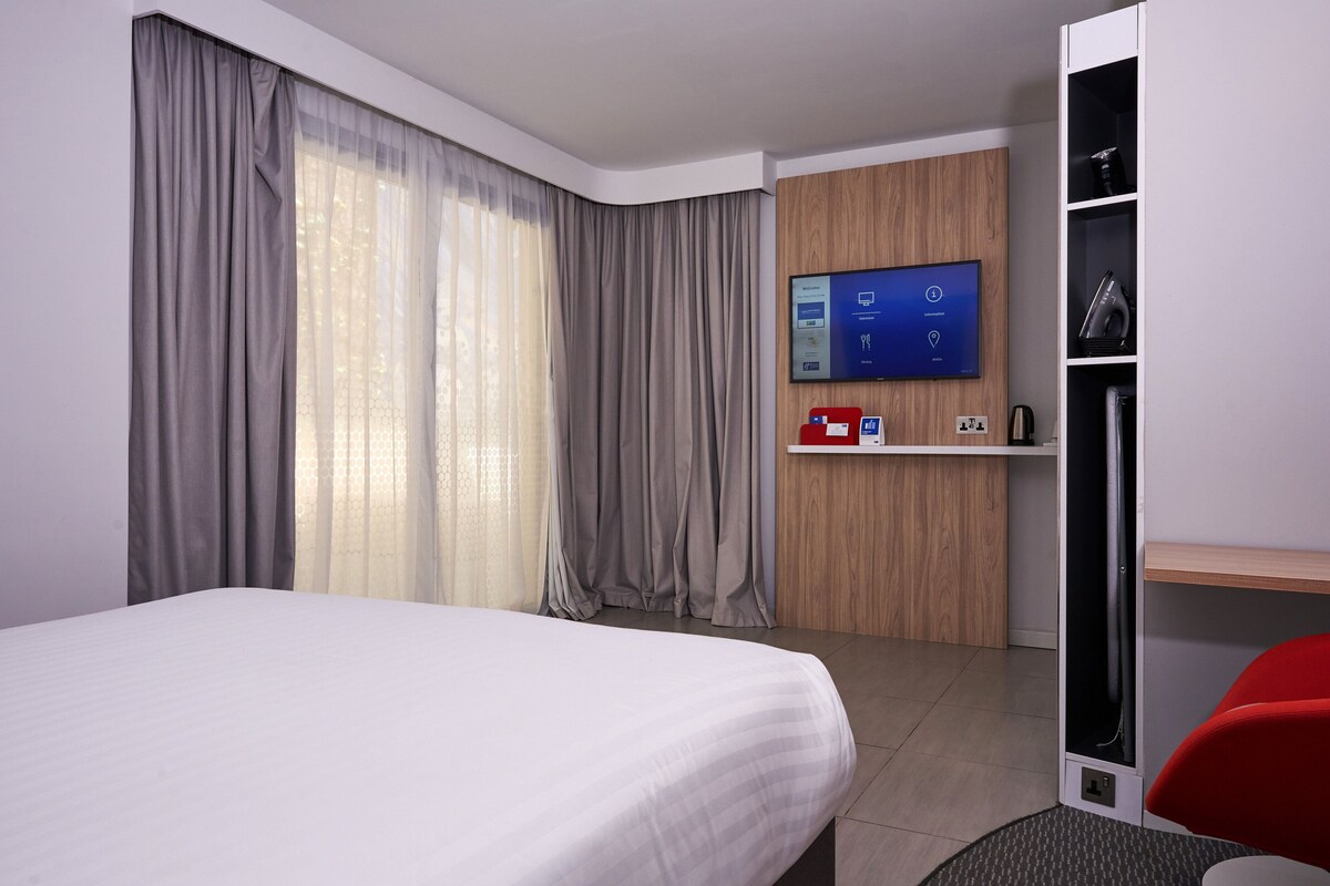A modern hotel room with a large bed, a TV mounted on the wall, a desk with a computer, and a wardrobe.