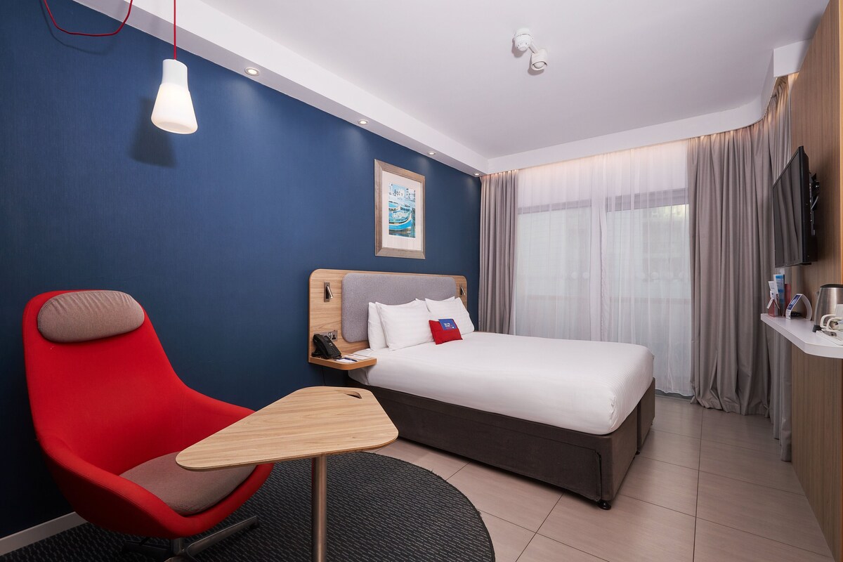 Una camera d'albergo con un letto, una sedia rossa, un tavolino, una TV e una finestra con tende.