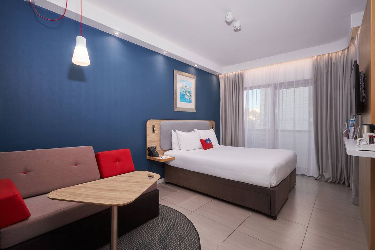 A hotel room with a bed, a sofa, a small table, a desk, a TV, and a window with curtains.