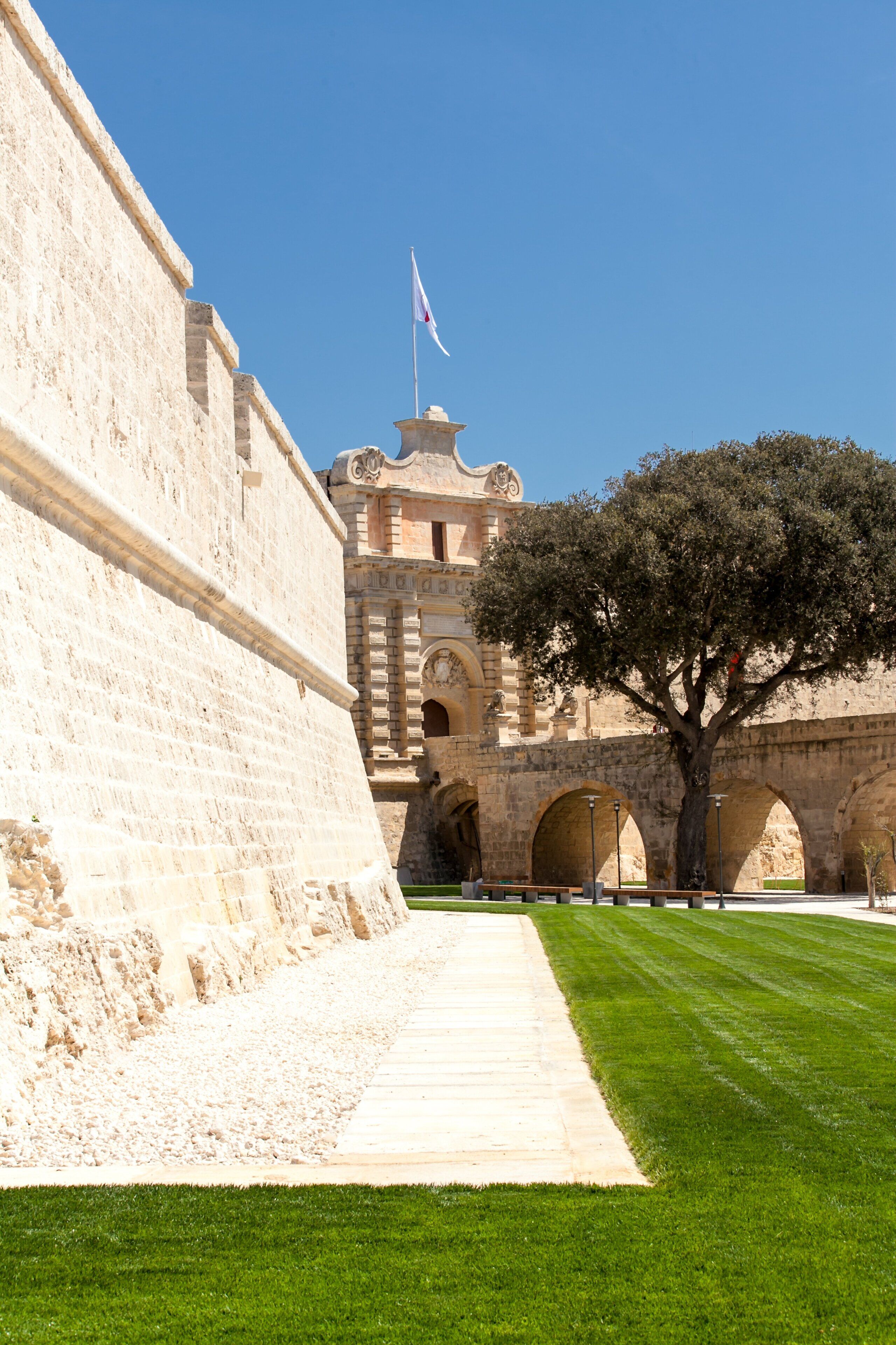 A historic stone building with a flag, a tree, and a paved walkway.