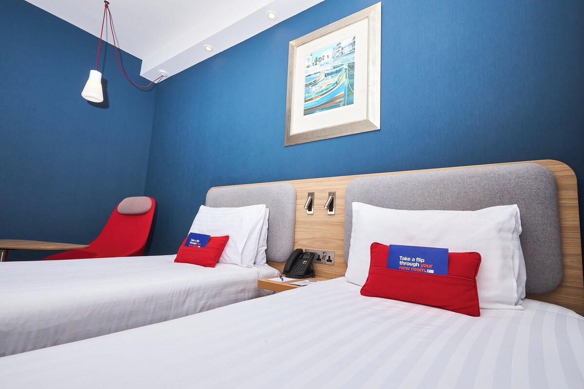 A hotel room with two beds, a red chair, a desk, and a framed picture on the wall.
