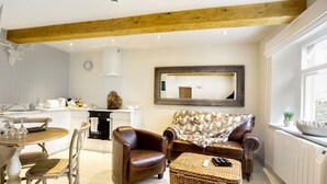 A cozy living room with a wooden ceiling, a dining area with a table and chairs, a brown leather sofa, a woven ottoman, and a large mirror on the wall. - Linton Court Coach House (Settle)