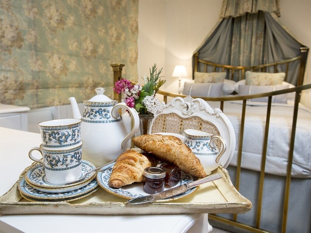 Bandeja de desayuno con juego de té, croissants y mermelada sobre una mesa en una habitación con cama y cortinas.