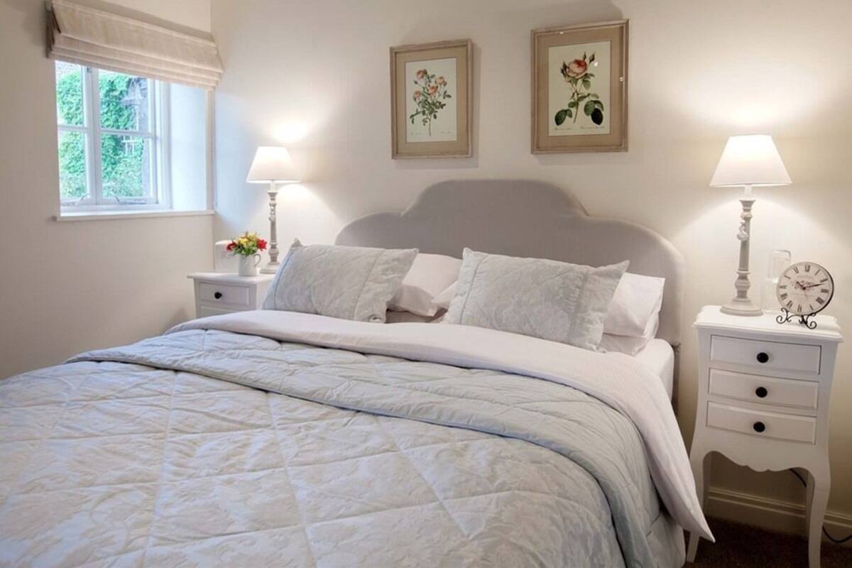 Une chambre à coucher comprenant un lit, deux tables de chevet, une fenêtre avec des rideaux et deux reproductions botaniques encadrées accrochées au mur.