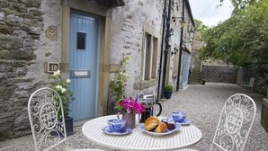 A stone building with a blue door, a table with white chairs, and a small garden area with flowers and a coffee pot. - Linton Court Coach House (Settle)
