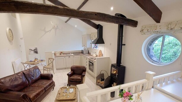 A cozy living room with a brown leather sofa, a white dining table, and a kitchen area with a stove and window. - Linton Court Coach House (Settle)