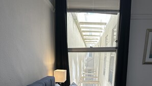 A neatly made bed with white linens and pillows, a bedside lamp, and a view of a cityscape through a window with dark curtains. - Hotel Ikon (San Francisco)
