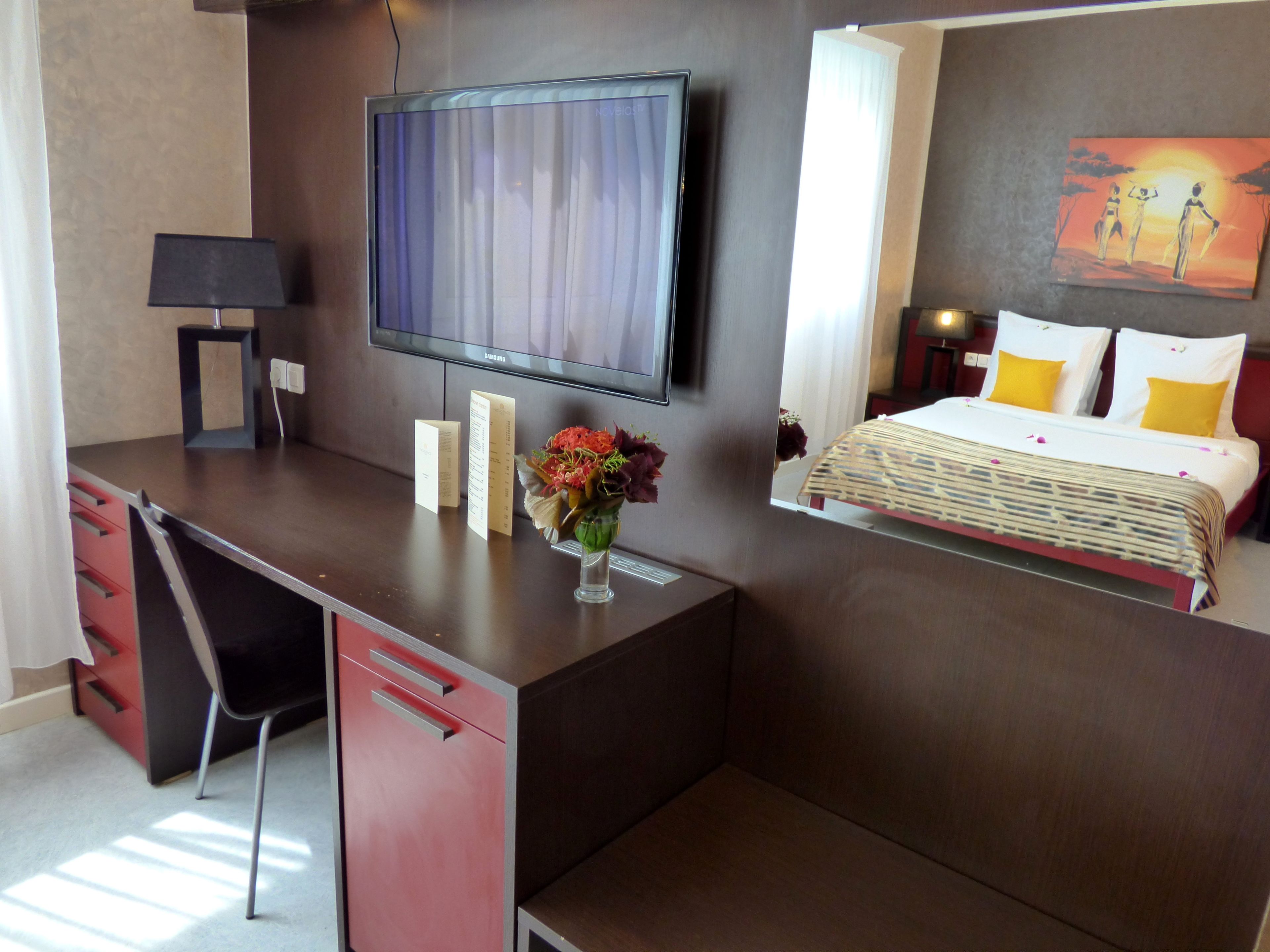 Habitación de hotel con una televisión de pantalla plana montada en la pared, un escritorio con una silla, una cama con colcha blanca y amarilla, y un cuadro en la pared.