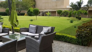 Un área de estar al aire libre con mobiliario de mimbre y una mesa de vidrio, rodeada de un césped cuidado y diversas plantas.