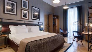 A hotel room with a large bed, a desk, a chair, a wardrobe, and framed pictures on the wall.