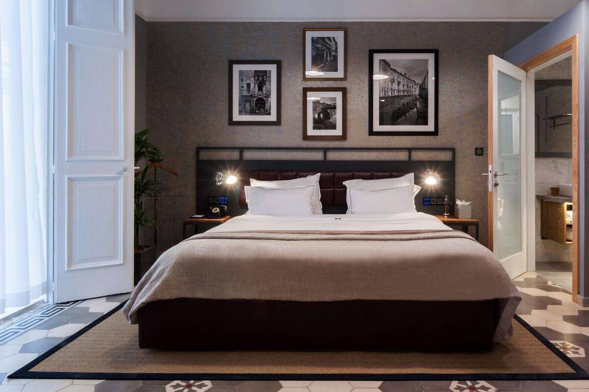 Ein modernes Hotelzimmer mit einem großen Bett, einer Kopfteilbeleuchtung, einem Nachttisch mit Lampe und gerahmten Bildern an der Wand.