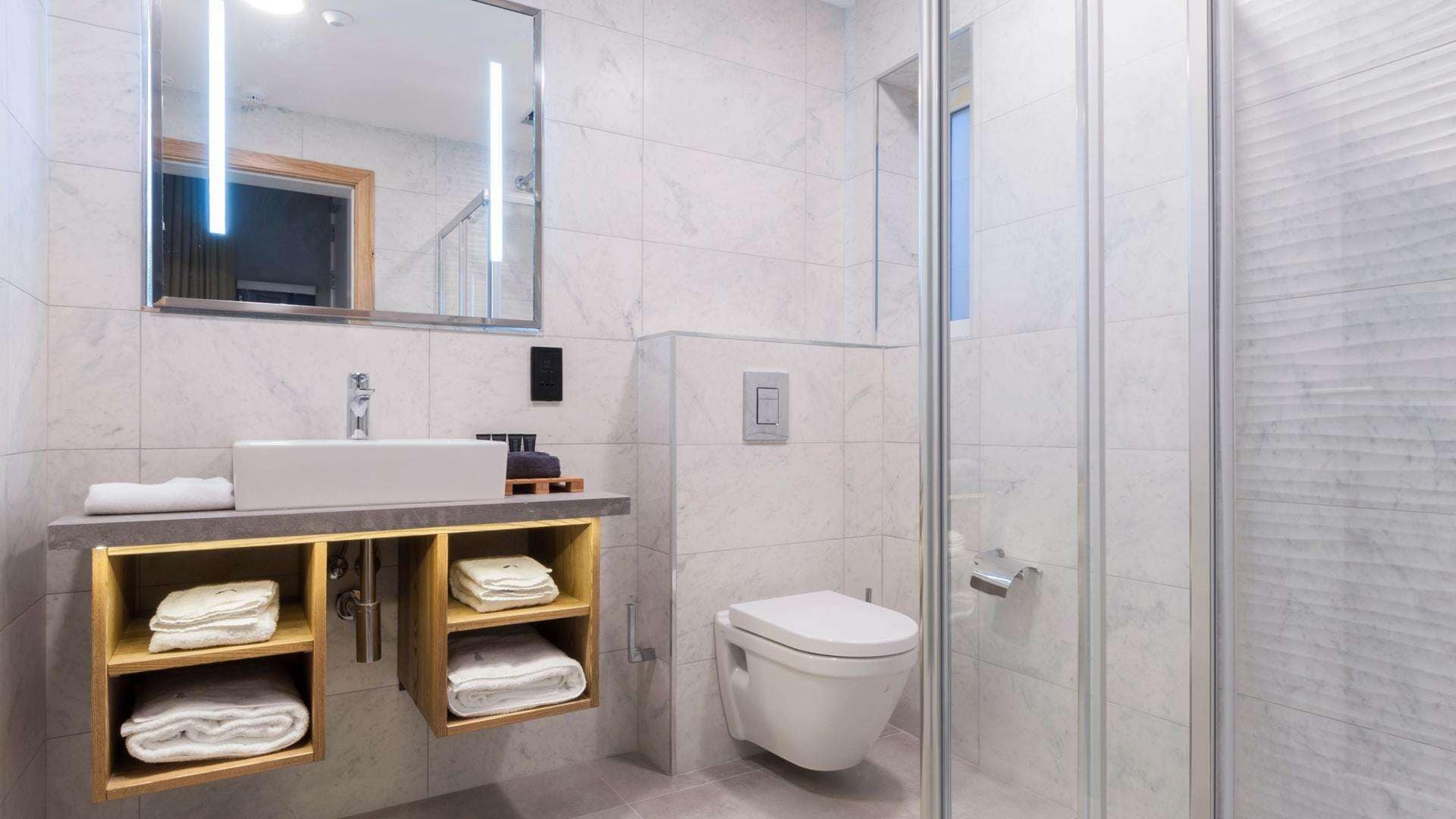 A modern bathroom with a marble sink, a glass shower enclosure, and wooden shelves with towels.