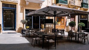 Un café al aire libre con mesas y sillas bajo sombrillas, la entrada de un edificio con un letrero y una puerta con un símbolo de cruz.