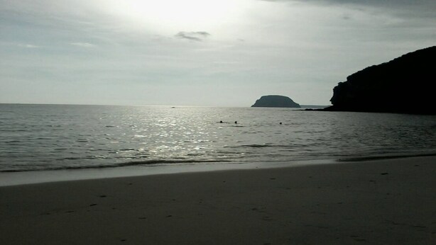 Ein Strand mit ruhiger See, einer entfernten Insel und einer felsigen Klippe.