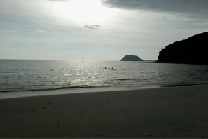 A beach with a calm sea, a distant island, and a rocky cliff.