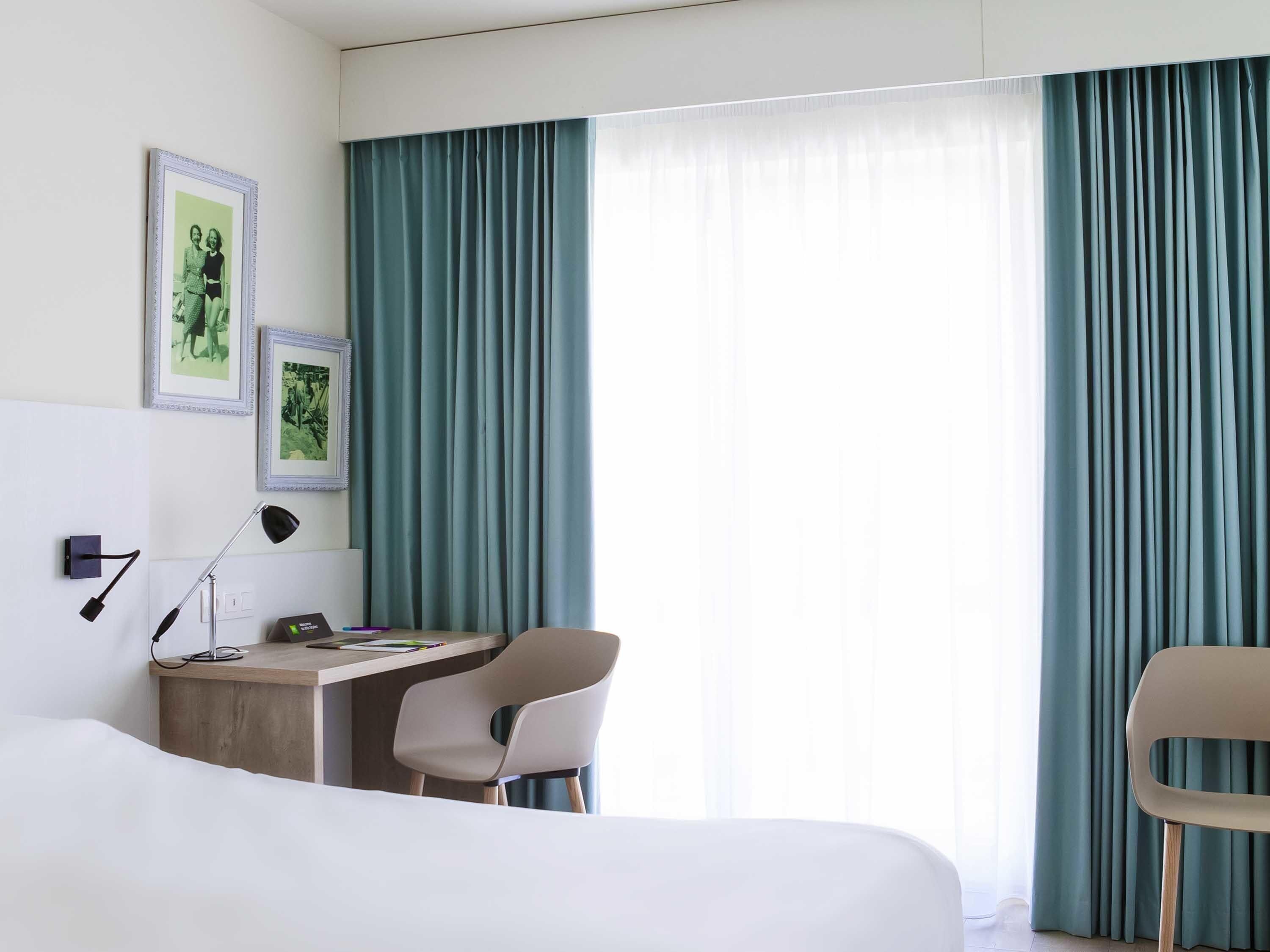 Habitación de hotel con cama, escritorio con silla, lámpara y dos cuadros en la pared.
