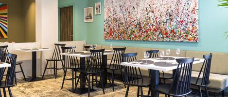 Un espace repas avec des tables et des chaises, une grande peinture abstraite au mur et une plante sur la droite.