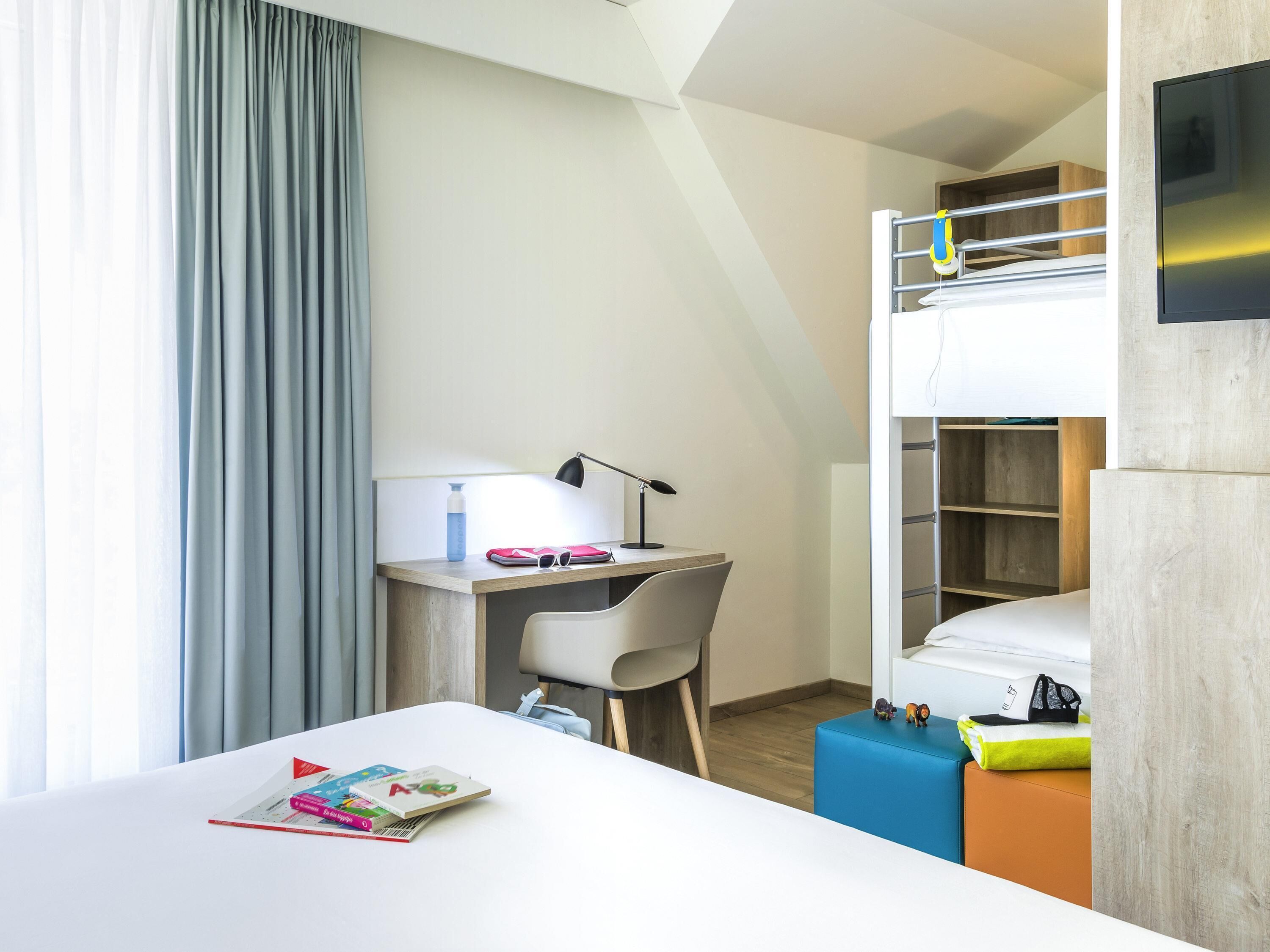 A modern hotel room with a bunk bed, a desk with a chair, a small table with a lamp, and a bed with a pillow and a blanket.