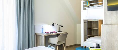 A modern hotel room with a bunk bed, a desk with a chair, a small table with a lamp, and a bed with a pillow and a blanket.