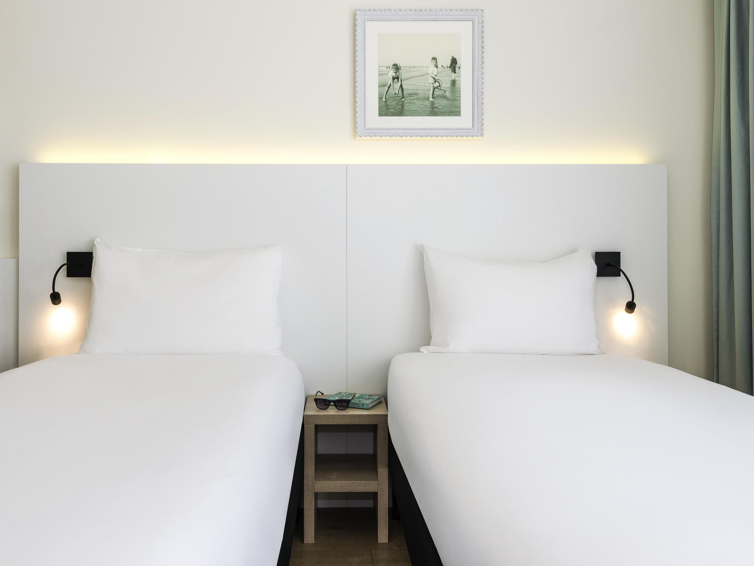 Een hotelkamer met twee eenpersoonsbedden, een klein nachtkastje met lamp en een lijst met een afbeelding aan de muur.
